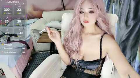 Video of a young Asian woman with long, wavy, pastel pink hair, wearing a black lace bra, sitting on a white couch in a cluttered room.