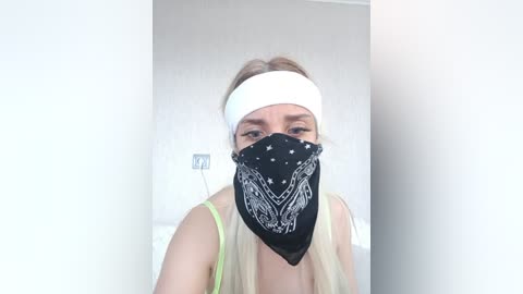 A video of a woman with light skin, wearing a white headband and a black bandana mask with white floral patterns. She has light brown hair tied back, and a green tank top. The background is plain white.