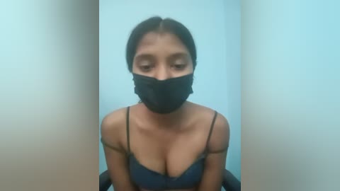 A video of a young woman with medium skin tone, dark hair, and wearing a black face mask, black spaghetti strap top, and sitting against a plain white background.