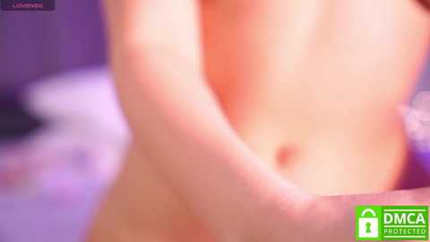 A blurred video of a nude, fair-skinned person, possibly female, with small breasts, resting their hand on their abdomen, in a softly lit bedroom with purple and white bedding.