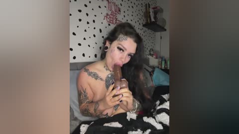 Video of a tattooed woman with dark hair, topless, performing oral sex on a large, circumcised penis in a bedroom with a polka dot wall and scattered belongings.