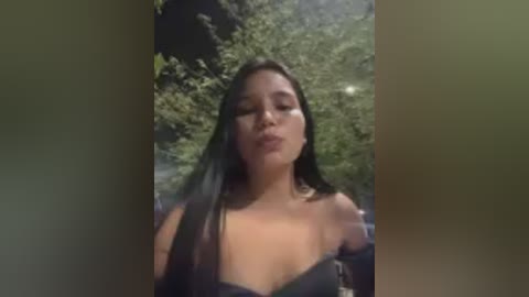 Video of a young Asian woman with long black hair, wearing a black off-shoulder top, standing outdoors against a blurred green background.