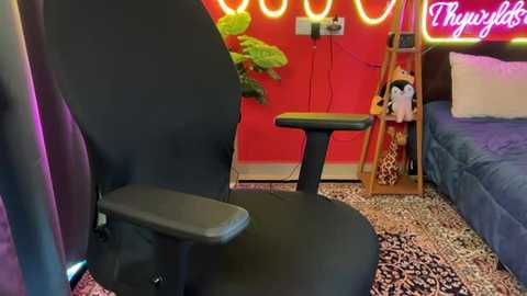 A video of a modern gaming room with a black gaming chair, leopard-print rug, blue bed, red wall, green plant, and \"Chuppers\" neon sign.