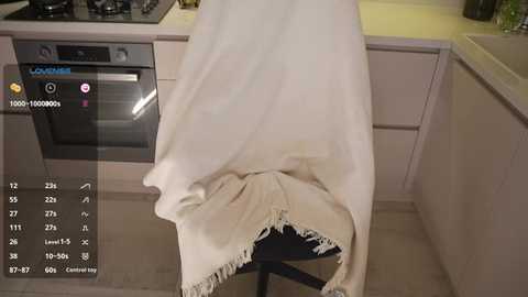 Video of a beige blanket draped over a black stool in a modern kitchen with light-colored cabinets and a stainless-steel oven. The image includes a virtual thermometer overlay.
