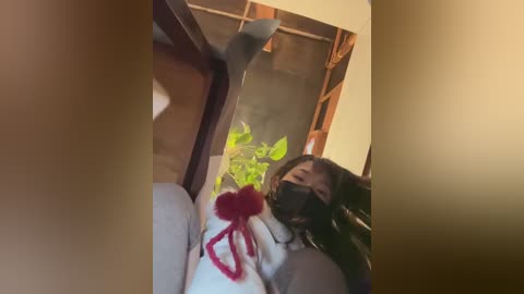 A candid video of a woman with long dark hair, wearing a face mask, lying on a wooden floor. She's dressed in a white top and has a red ribbon tied to her left hand. The background includes a green plant and wooden furniture.