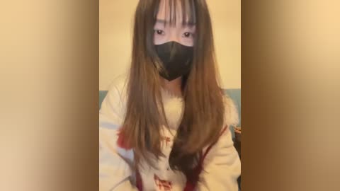 Video of an Asian woman with long brown hair, wearing a black face mask, white long-sleeved shirt, and sitting on a couch against a plain beige wall background.