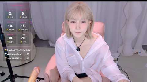Video of a young, light-skinned Asian woman with short blonde hair, wearing a white hoodie and necklace, sitting in a pink chair. A man's hand holds a dildo in front of her. Background includes a white curtain and a screen with streaming statistics.