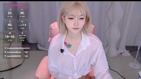 A video of a young Asian woman with shoulder-length blonde hair, wearing a sheer white blouse, sitting on a pink chair in a dimly lit room with white curtains.