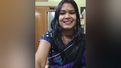 Video of a smiling woman with medium skin tone, long black hair, and a red bindi, wearing a black sari with purple and white patterns. She is indoors, with a kitchen background visible.