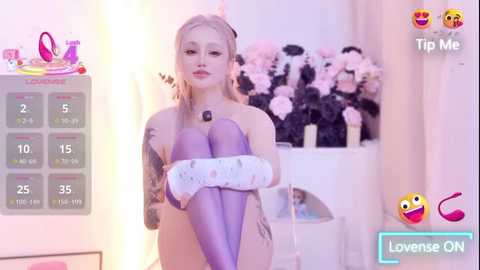 Video of a blonde, tattooed woman in a purple, sheer bodysuit, sitting on a white stool in a brightly lit room with pink flowers and a calendar in the background.