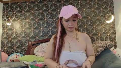 Video of a young, fair-skinned woman with long pink hair, wearing a pink baseball cap and white tank top, sitting on a bed with a geometric-patterned wallpaper, surrounded by stuffed toys and a green pillow.