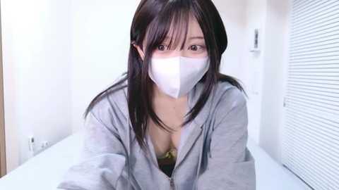 Video of an Asian woman with long black hair, wearing a white face mask, grey hoodie, and revealing a yellow bra underneath, in a minimalist white bathroom setting.