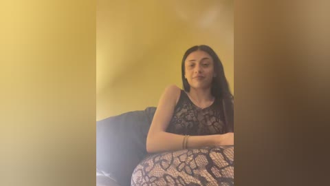 Video of a young woman with long, dark hair, wearing a black lace dress, sitting on a black couch against a mustard-yellow wall. Her expression is neutral.