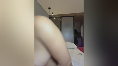 Video of a nude person's shoulder and arm, with blurred face, standing in a modern bedroom with a bed, wardrobe, and wooden wall.