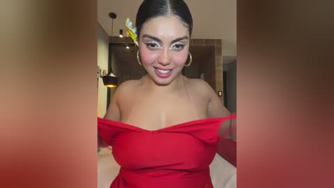 Video of a smiling Latina woman with medium skin tone, dark hair pulled back, wearing a red off-shoulder dress, large hoop earrings, in a modern, dimly-lit bedroom with a bed, wall art, and a hanging lamp.