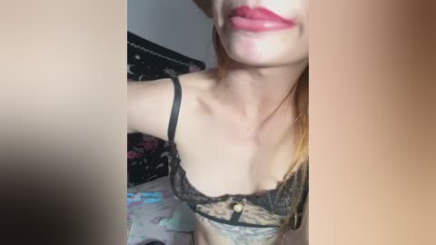 Video of a fair-skinned woman with long, straight red hair, wearing a black lace bra, smiling with full, pink lips. Background features a dark, patterned blanket and a colorful, floral-patterned sheet.