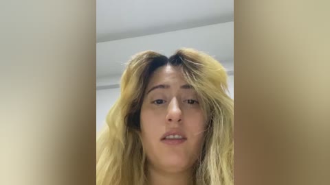 A video of a young woman with long, tousled blonde hair and light skin, looking slightly confused. She has a neutral expression and is indoors, with white walls and a ceiling visible in the background.