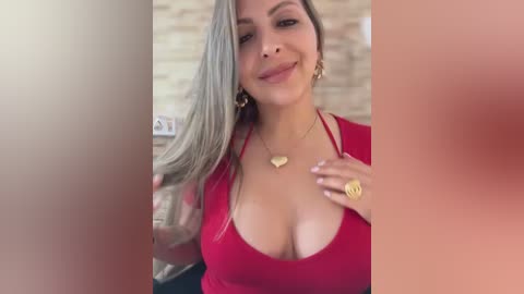 Video of a smiling, light-skinned woman with long, straight, silver hair, wearing a low-cut red dress, gold jewelry, and a heart pendant necklace. Background shows a blurred, beige stone wall.