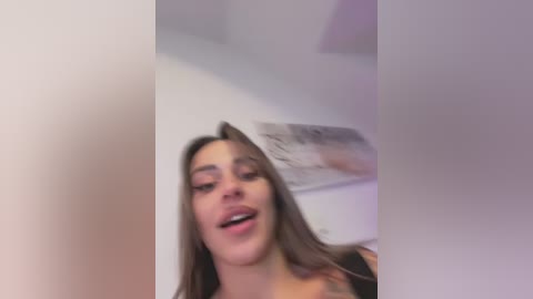 A blurry video of a young woman with light brown hair and a slight smile, captured in an indoor setting with white walls and a framed photo in the background.