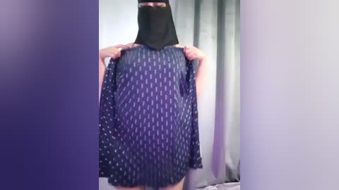 A video of a person in a black face mask, holding a blue shirt with white dots, against a backdrop of gray curtains.