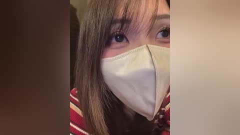 A close-up video of a woman with light skin and straight brown hair, wearing a beige medical mask and a red-striped sweater. Her brown eyes are partially visible. The background is blurred.