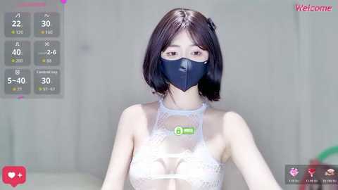 A video of an East Asian woman with short black hair, wearing a white lace bra, blue mask, and blue gloves, in a sterile, white-lit room with digital health stats.