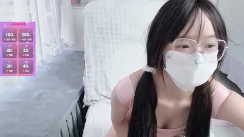 Video of an Asian woman with long black pigtails and glasses wearing a white mask, seated on a white cushioned chair in a minimalist room with a grey floor and white furniture.