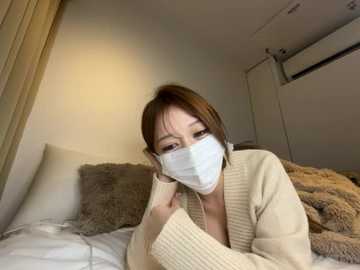 A video of an Asian woman with short brown hair, wearing a white face mask, beige cardigan, and glasses, lying on a bed with beige and brown blankets, in a cozy, dimly lit room.