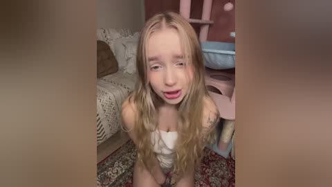Video of a young blonde girl with long wavy hair, wearing a white tank top, kneeling on a patterned rug in a cozy, beige-painted room with a cat scratching post in the background.