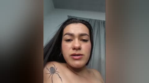 A video of a Latina woman with medium skin tone, long black hair, and a spider tattoo on her left shoulder. She has a neutral expression and is indoors, with grey curtains in the background.
