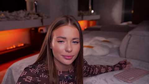 Video of a young Caucasian woman with straight brown hair, light skin, and subtle makeup, wearing a black patterned sweater, sitting on a white couch in a dimly lit living room with a fireplace and a laptop on the table.