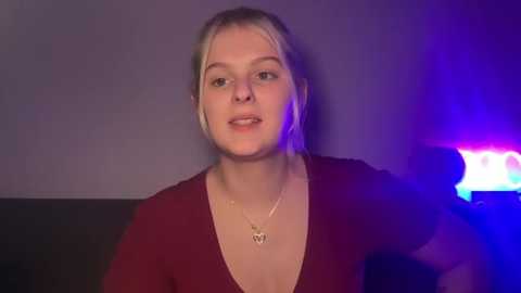 Video of a young white woman with blonde hair, wearing a deep burgundy V-neck top, smiling slightly, standing against a dimly lit purple backdrop, with a blue light source illuminating her face.
