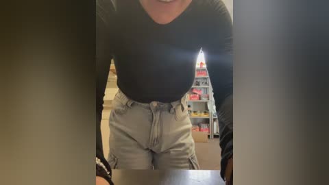 Video of a woman with light skin, wearing a black long-sleeve top and high-waisted light gray cargo pants, standing in a cluttered room with a bookshelf and toys in the background.