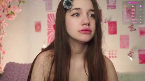 Video of a young woman with long brown hair, blue flower headpiece, and nose ring, in a pink and white bedroom with floral decor and video game controller.