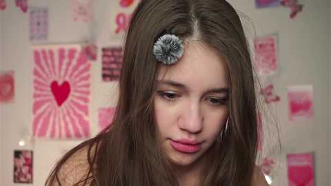 Video of a young girl with straight brown hair, adorned with a gray flower clip, wearing a pink top, against a pink and white heart-decorated wall.