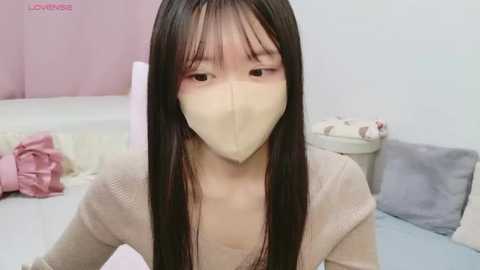 Video of an Asian woman with long black hair, wearing a beige face mask and a beige cardigan, sitting on a bed with pink and white bedding.