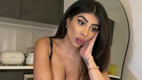 Media: Video of a Latina woman with long black hair, large brown eyes, and dark eyebrows, wearing a black bra, posing seductively in a modern kitchen.