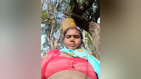 A video of a woman wearing a vibrant pink sari with a blue shawl, her head adorned with a yellow pot, standing in a forest with tall trees and sunlight filtering through.