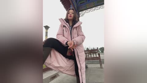 Video of a woman with medium-length brown hair, wearing a pink puffy coat and black pants, sitting on a stone bench in a park, under a red and blue tiled pavilion.