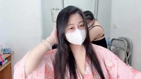 Video of a young Asian woman with long black hair, wearing a pink robe with floral patterns, a white face mask, and a white headband. She's in a simple, white-walled room with a mirror and a metal chair in the background.