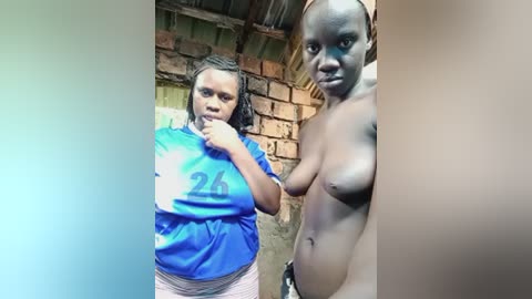 A video shows two African women standing in a rustic, brick-walled room. One wears a blue jersey with the number \"26\" and the other is topless, revealing her large breasts.