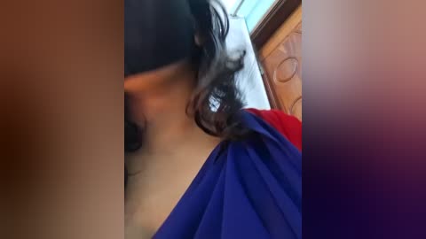 A blurry video shows a person in a black mask, wearing a red and blue outfit, with wavy dark hair. The background features a wooden door and wall.