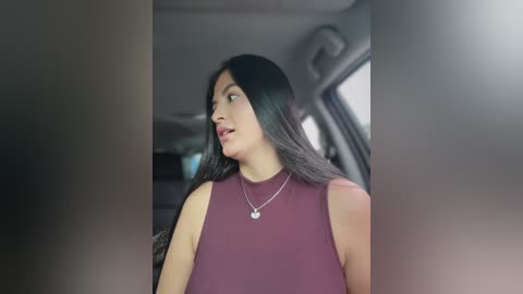 A video captures a woman with long, straight black hair, wearing a sleeveless maroon top, seated in a car with blurry, muted background. She has a light skin tone and a neutral expression, with a silver necklace.