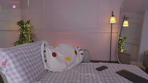Video of a cozy bedroom with a white duvet featuring cartoon cat pattern, checkered pillow, and a nightstand with a lamp and fairy lights.