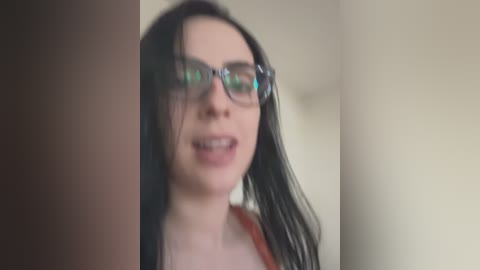 A blurred video of a woman with long black hair, wearing glasses, and a red top, standing against a beige wall.