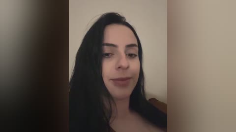 Video of a young woman with long, straight black hair, fair skin, and a slight smile, standing against a beige wall.