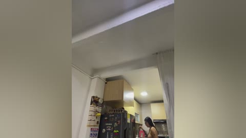 Video of a small, dimly lit kitchen with white walls, a refrigerator, and a woman in a sleeveless top and black pants. The image is slightly blurred, possibly due to motion.