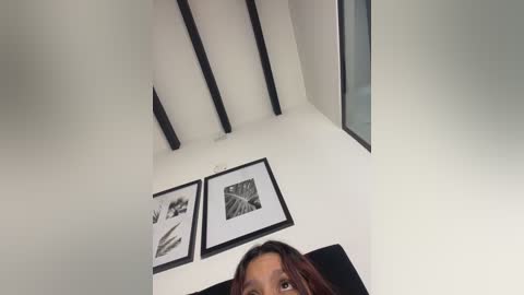 Video of a woman with medium brown skin and long, wavy brown hair, looking up at a modern, minimalist room with white walls, black beams, and black-and-white framed artwork on the wall.