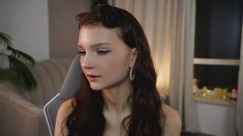 Video of a young woman with fair skin, long dark hair, and delicate features, wearing a white top, seated indoors, looking down. Background features a beige sofa, potted plant, and dimly lit room with curtains.