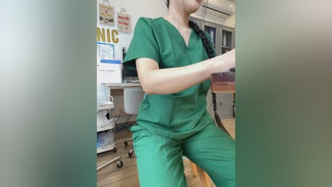 Video of a female nurse in green scrubs with a black braid, standing in a medical office with white walls, a desk, and posters.